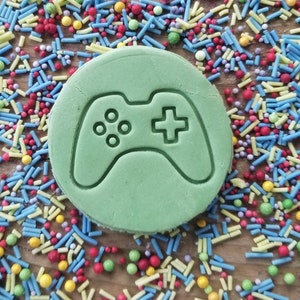 May include: A circular, pale green biscuit featuring a video game controller design. The biscuit is surrounded by colourful sprinkles in blue, yellow, red, and green, on a wooden surface. A playful treat for gamers.