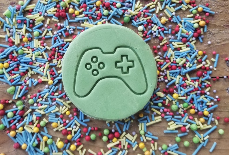 May include: A circular, pale green biscuit featuring a video game controller design. The biscuit is surrounded by colourful sprinkles in blue, yellow, red, and green, on a wooden surface. A playful treat for gamers.