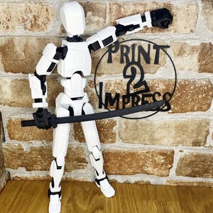 May include: A white articulated action figure with black detailing, holding a black sword. The figure is posed dynamically against a brick wall. The figure has a robotic design. The text "PRINT 2 IMPRESS" is visible in the background.