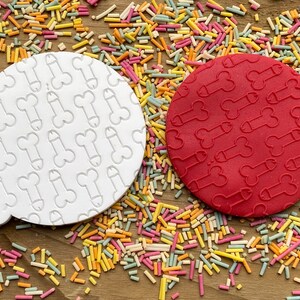 May include: Two round cookie cutters, one white and one red, each imprinted with a repeating phallic design. The cutters are surrounded by colorful sprinkles on a wooden surface. The white cutter has a small handle.