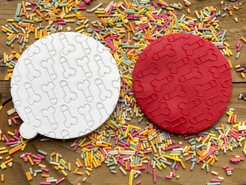 May include: Two round cookie cutters, one white and one red, each imprinted with a repeating phallic design. The cutters are surrounded by colorful sprinkles on a wooden surface. The white cutter has a small handle.