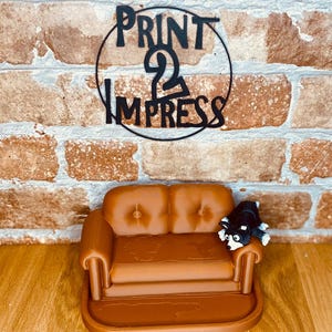 May include: A small brown sofa with a black and white dog ornament. Above the sofa is a black metal sign with the words "PRINT 2 IMPRESS" in a circular design. The background is a brick wall.