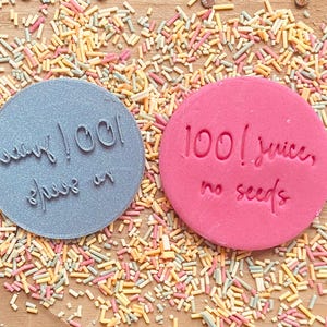 May include: Two round, pink and gray silicone stamps with text. The pink stamp says "100! juice, no seeds". The gray stamp says "nearly 100!  shh, it's a secret".