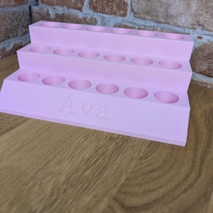 May include: A tiered, light pink organizer with multiple circular openings. The word "Ava" is engraved on the front. The organizer is made of a solid material and is designed to hold small items. It sits on a wooden surface.