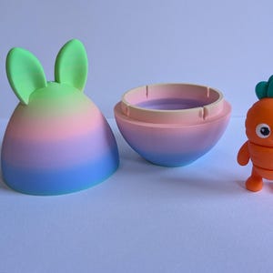 May include: A pastel rainbow colored egg-shaped container with bunny ears opens to reveal a small orange carrot character with a green leafy top and a smiling face.