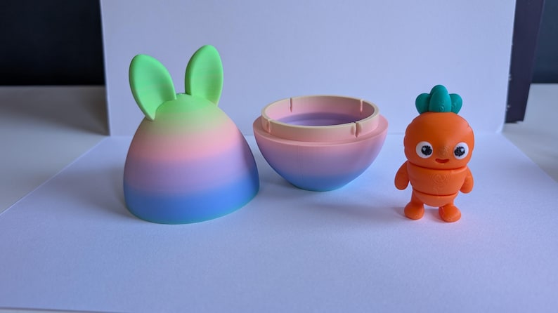 May include: A pastel rainbow colored egg-shaped container with bunny ears opens to reveal a small orange carrot character with a green leafy top and a smiling face.
