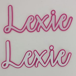May include: Two pink and white layered paper cutouts of the name "Lexie" in a cursive font.