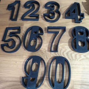 May include: Black, three-dimensional numbers 1 through 0. The numbers are in a sans-serif font and appear to be made of a textured material. The numbers are arranged in two rows on a wooden surface.