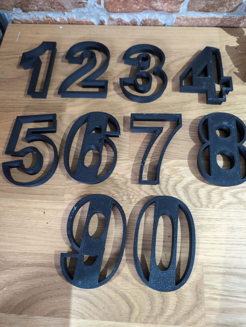 May include: Black, three-dimensional numbers 1 through 0. The numbers are in a sans-serif font and appear to be made of a textured material. The numbers are arranged in two rows on a wooden surface.