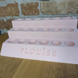 May include: A tiered, light pink makeup organizer with multiple circular compartments. The organizer has three levels and features the name "PLouise" embossed on the front. The item is made of a solid material and sits on a wooden surface.