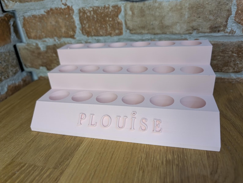 May include: A tiered, light pink makeup organizer with multiple circular compartments. The organizer has three levels and features the name "PLouise" embossed on the front. The item is made of a solid material and sits on a wooden surface.