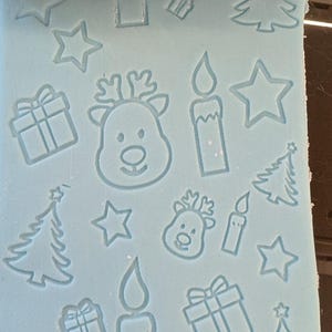 May include: A red rolling pin with Christmas-themed designs pressed into light blue dough. The designs include reindeer, candles, gift boxes, Christmas trees, and stars. The rolling pin is used to create festive patterns.