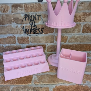 May include: A collection of pink 3D-printed organizers. Includes a tiered tray with holes, a crown-shaped stand, and a rectangular container, all with the name "PLOUISE" embossed. The background is a brick wall.