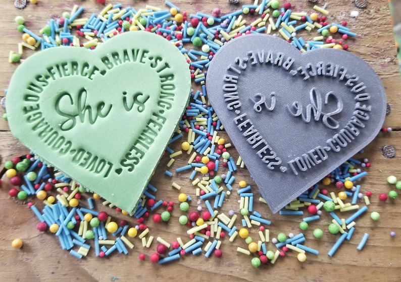 K&ouml;nnte beinhalten: Zwei herzf&ouml;rmige Stempel, einer gr&uuml;n und einer grau, mit den Worten "She is" in der Mitte. Der gr&uuml;ne Stempel hat die Worte "Loved, Courageous, Fierce, Brave, Strong, Fearless" am Rand. Der graue Stempel hat die gleichen Worte. Bunte Streusel umgeben die Stempel.