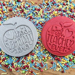 May include: Two circular cookie stamps, one grey and one red, featuring apple designs and the text "Best Teacher Ever." The stamps are surrounded by colourful sprinkles on a wooden surface.