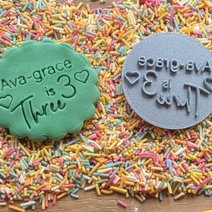 May include: A green cookie with the text "Ava-grace is 3 Three" and a heart shape, next to a gray stamp with the same text in reverse. The items are surrounded by colorful sprinkles.