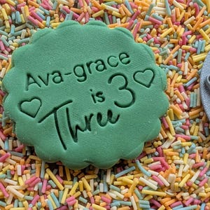 May include: A green, scalloped-edge cookie cutter with the text "Ava-grace is 3 Three" and heart shapes. The cutter is surrounded by colorful sprinkles. A gray circular stamp with the text "Ava-grace is 3" is also visible.