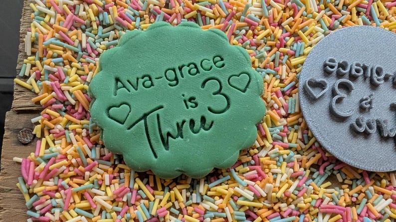 May include: A green, scalloped-edge cookie cutter with the text "Ava-grace is 3 Three" and heart shapes. The cutter is surrounded by colorful sprinkles. A gray circular stamp with the text "Ava-grace is 3" is also visible.