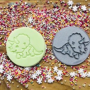 May include: Two circular cookie cutters with a dinosaur design. One is light green, the other is gray. The cutters are surrounded by colorful sprinkles and snowflake-shaped decorations on a wooden surface.