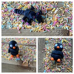 May include: Three black toys on a wooden surface covered in colorful sprinkles. The toys are a cat, a penguin, and another penguin. The cat is curled up and has a yellow eye. The penguins are standing upright and have orange feet.