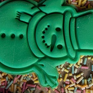 Op de afbeelding: Een groene koekjesvorm in de vorm van een sneeuwpop met hoed, sjaal en gezichtsdetails. De vorm is omgeven door kleurrijke hagelslag op een houten oppervlak. Perfect voor feestelijk bakken.