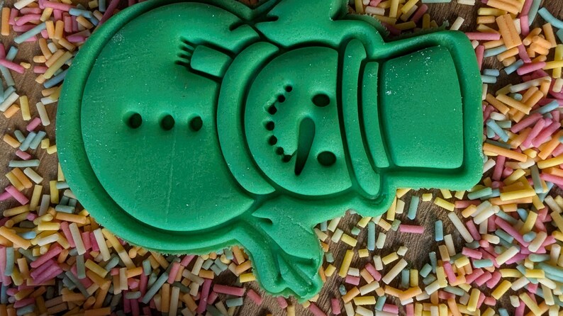 Op de afbeelding: Een groene koekjesvorm in de vorm van een sneeuwpop met hoed, sjaal en gezichtsdetails. De vorm is omgeven door kleurrijke hagelslag op een houten oppervlak. Perfect voor feestelijk bakken.