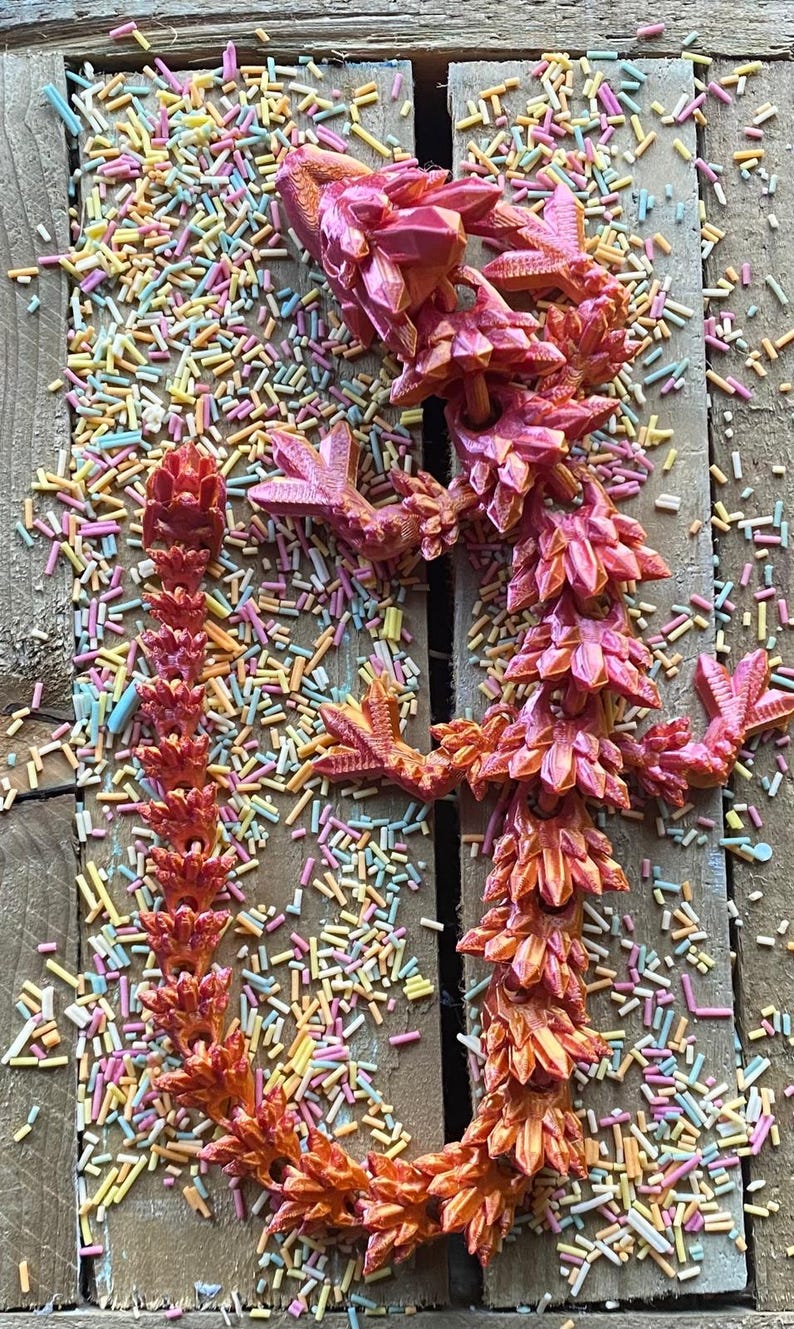 May include: A bright pink and orange 3D printed dragon figurine with a spiky, crystal-like design. The dragon is posed in a standing position with its wings spread out. The figurine is surrounded by colorful sprinkles.