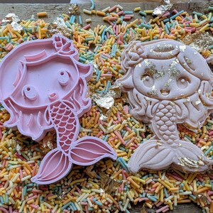 May include: Two mermaid-shaped cookie cutters, one pink and one decorated with gold leaf, rest on a bed of colorful sprinkles. The pink cutter is a clean outline, while the other is a finished cookie.