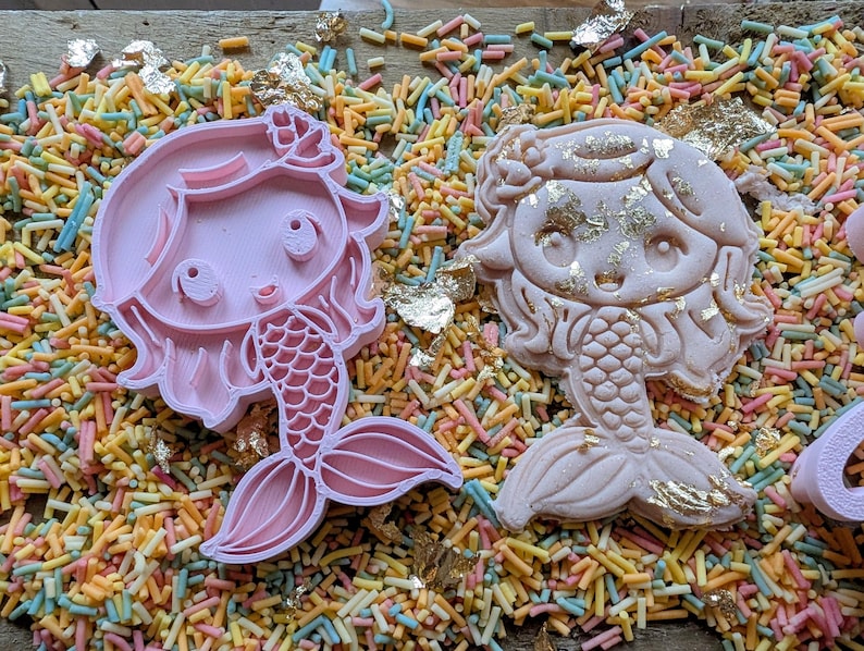 May include: Two mermaid-shaped cookie cutters, one pink and one decorated with gold leaf, rest on a bed of colorful sprinkles. The pink cutter is a clean outline, while the other is a finished cookie.