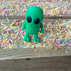 K&ouml;nnte beinhalten: Eine gr&uuml;ne Alien-Figur mit schwarzen Augen steht auf einer Holzoberfl&auml;che, die mit bunten Streuseln bedeckt ist.