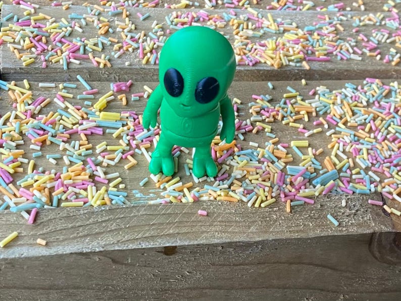 K&ouml;nnte beinhalten: Eine gr&uuml;ne Alien-Figur mit schwarzen Augen steht auf einer Holzoberfl&auml;che, die mit bunten Streuseln bedeckt ist.