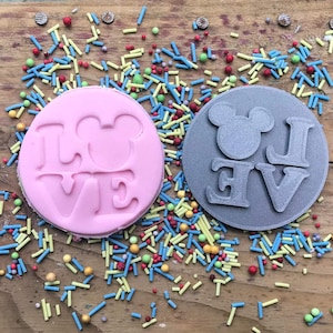May include: A pink, circular biscuit with the word "LOVE" embossed, featuring a Mickey Mouse head as the "O". Beside it is a grey biscuit stamp with the same design. The biscuits are on a wooden surface scattered with colourful sprinkles.