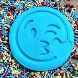 May include: A circular, bright blue emoji-shaped biscuit featuring a winking face and a heart. The biscuit is set amongst a vibrant array of sprinkles in red, blue, yellow, and green, creating a colourful and fun display.