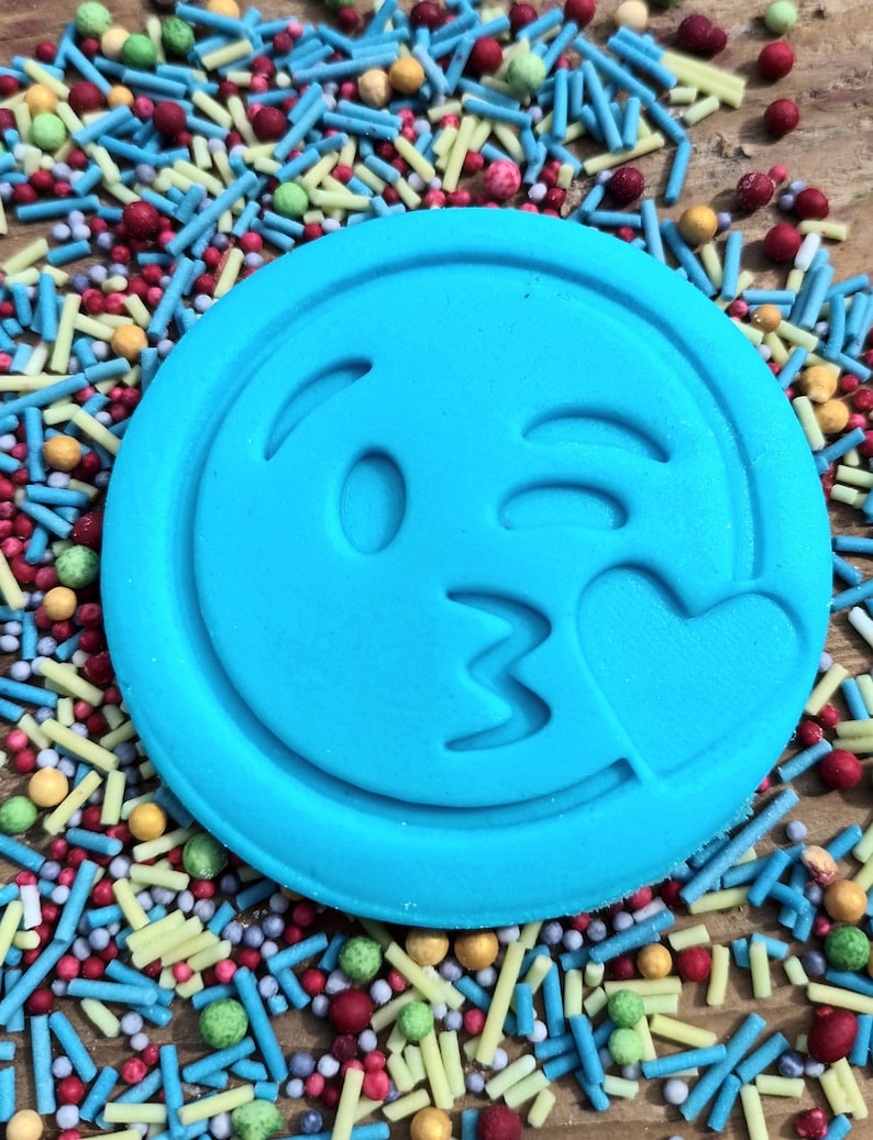 May include: A circular, bright blue emoji-shaped biscuit featuring a winking face and a heart. The biscuit is set amongst a vibrant array of sprinkles in red, blue, yellow, and green, creating a colourful and fun display.