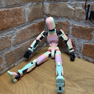 May include: A poseable artist's mannequin in pastel colors, including pink, blue, and yellow. The figure has articulated joints and is positioned on a wooden surface, with a brick wall in the background.