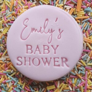 Op de afbeelding: Een ronde roze koek met de tekst "Emily's Baby Shower" in roze letters.