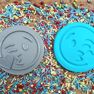 May include: Two circular cookie cutters, one grey and one blue, each depicting a winking emoji blowing a kiss. The cutters are surrounded by colourful sprinkles on a wooden surface, ideal for baking themed treats.
