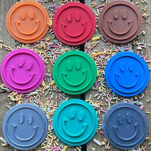 May include: Twelve round, colorful cookie cutters in the shape of a smiley face. The cutters are arranged in a circle on a wooden surface with colorful sprinkles.