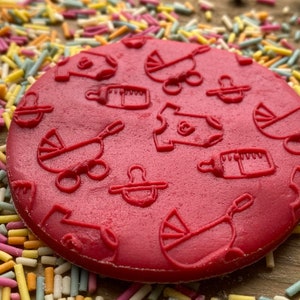 May include: Red silicone baby themed cookie stamp with designs of a baby bottle, a baby carriage, a pacifier, and a onesie.