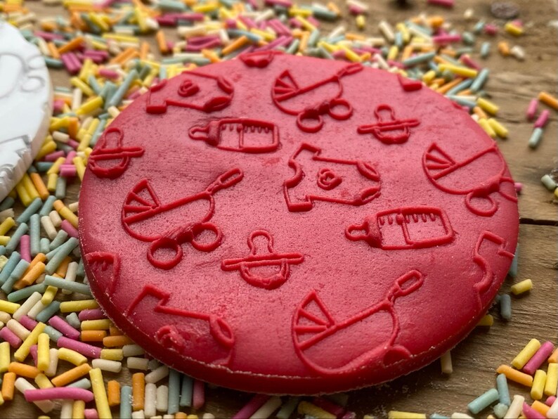 May include: Red silicone baby themed cookie stamp with designs of a baby bottle, a baby carriage, a pacifier, and a onesie.