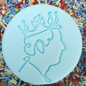 May include: A light blue, round cookie with a detailed outline of a crown and profile. The cookie is surrounded by colorful sprinkles. The design is a tribute to a royal figure.