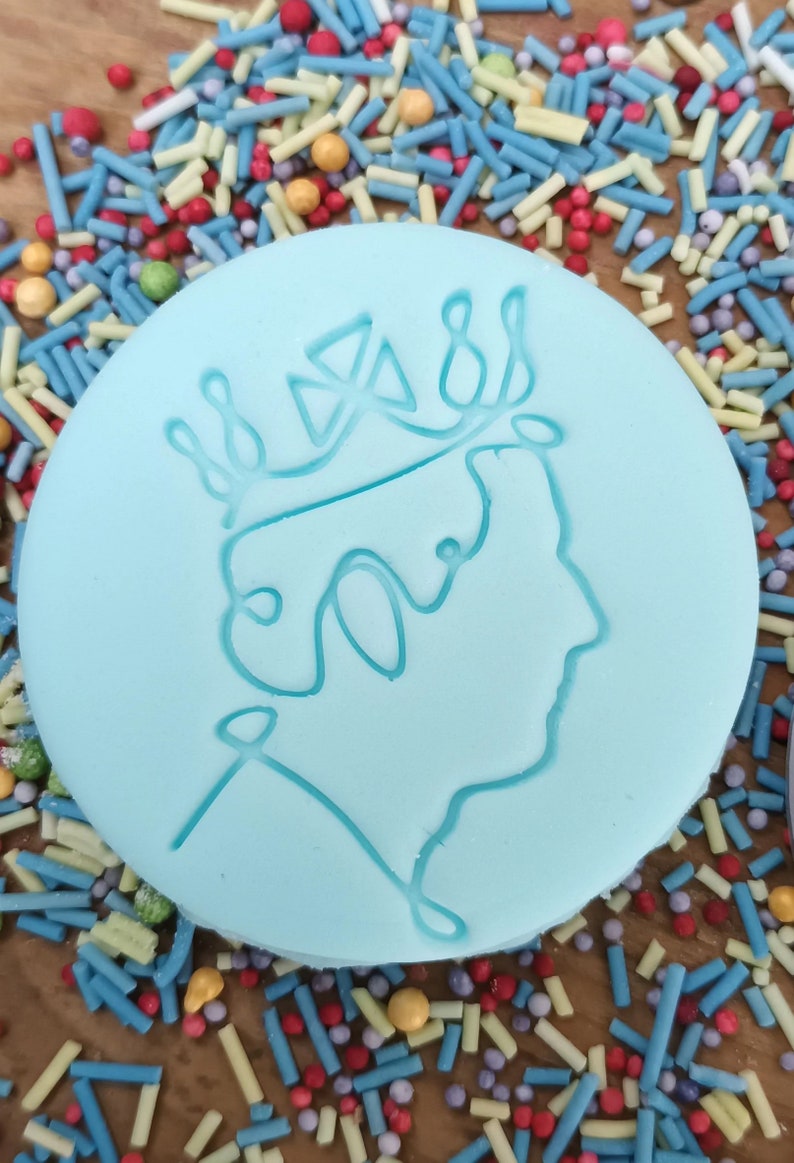 May include: A light blue, round cookie with a detailed outline of a crown and profile. The cookie is surrounded by colorful sprinkles. The design is a tribute to a royal figure.