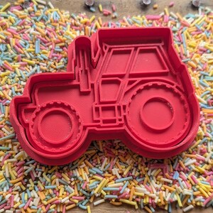 May include: A red plastic tractor-shaped cookie cutter. The cutter features the shape of the tractor's wheels, cab, and exhaust pipe. The cutter is surrounded by colourful sprinkles.