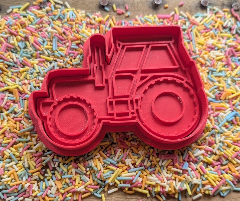 May include: A red plastic tractor-shaped cookie cutter. The cutter features the shape of the tractor's wheels, cab, and exhaust pipe. The cutter is surrounded by colourful sprinkles.