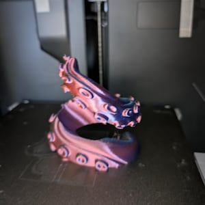 May include: A 3D-printed octopus tentacle sculpture in pink, purple, and blue hues. The tentacle has suction cups and a curved shape. It appears to be a decorative item or part of a larger artwork.
