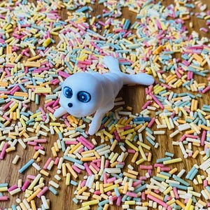 May include: A white ceramic seal figurine with blue eyes, lying on a surface covered in colorful sprinkles.