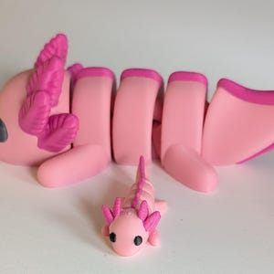 May include: Two pink axolotl figurines, one larger than the other. The larger axolotl has segmented body parts and pink frilly gills. The smaller axolotl is a solid piece.