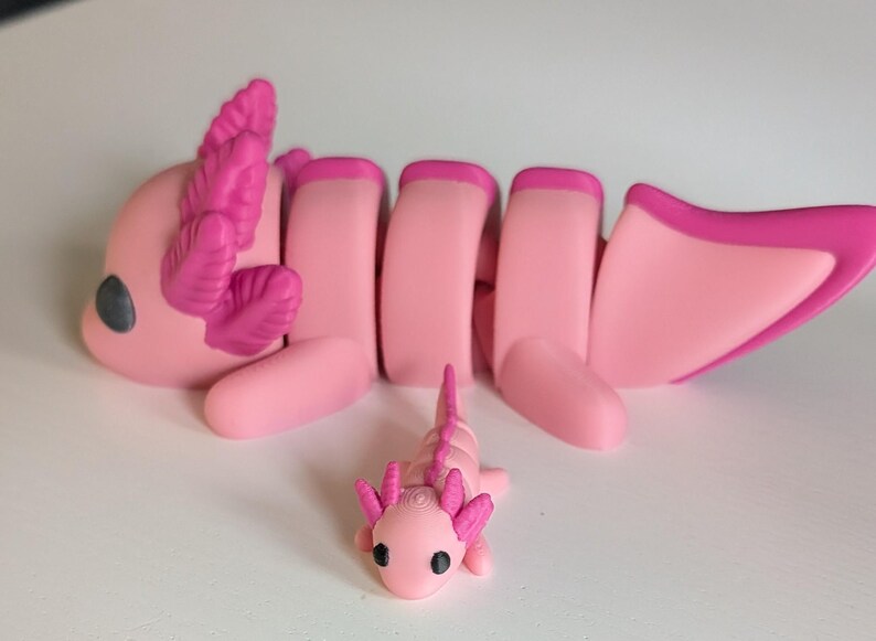 May include: Two pink axolotl figurines, one larger than the other. The larger axolotl has segmented body parts and pink frilly gills. The smaller axolotl is a solid piece.