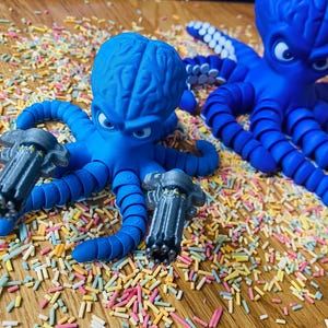 May include: Two blue octopus figurines with angry expressions and gray gun attachments. The octopus figures are surrounded by colorful sprinkles on a wooden surface. One octopus is holding two guns.