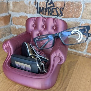 May include: A burgundy-colored miniature armchair-shaped organizer, holding a pair of black-framed eyeglasses, keys, and a small wallet. The chair has button tufting and curved armrests. The text "PRINT 2 IMPRESS" is visible in the background.